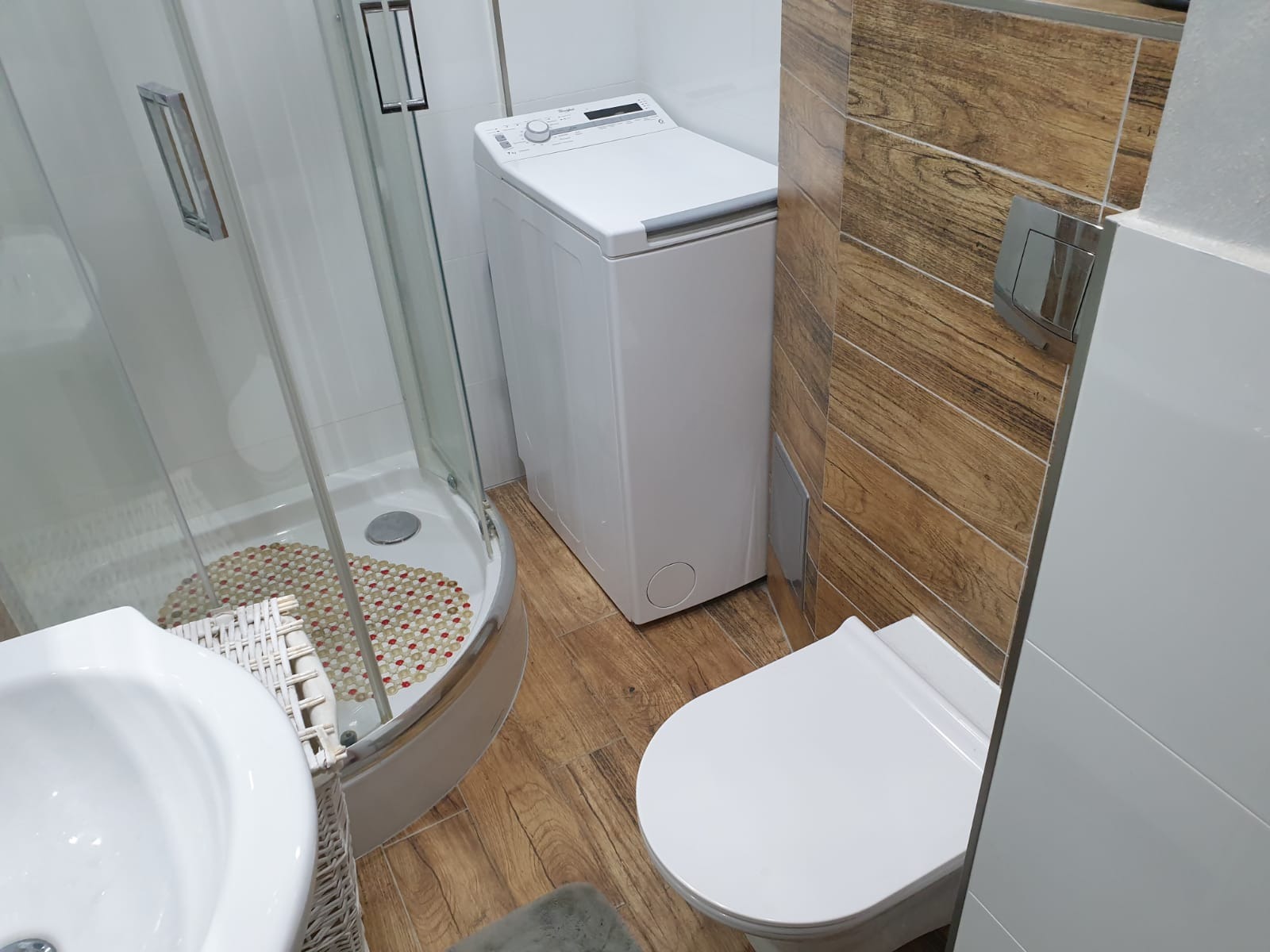Nowoczesna łazienka z kabiną prysznicową, pralką ładowaną od góry i toaletą, wykończona płytkami imitującymi drewno na jednej ze ścian.