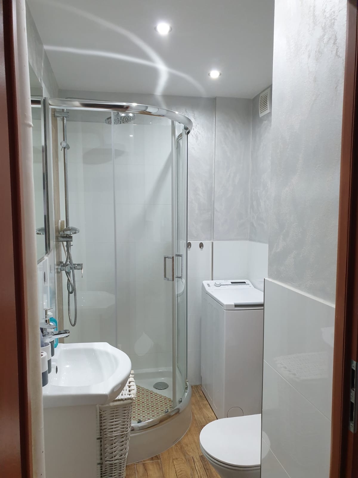 Funkcjonalna łazienka po remoncie: kabina prysznicowa z chromowanymi elementami, umywalka z szafką z wikliny, pralka i toaleta w jasnych barwach, ściany wykończone dekoracyjnym tynkiem.