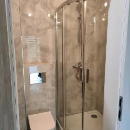 Wykończona łazienka z kabiną prysznicową narożną, wiszącą toaletą i płytkami imitującymi marmur na ścianach.