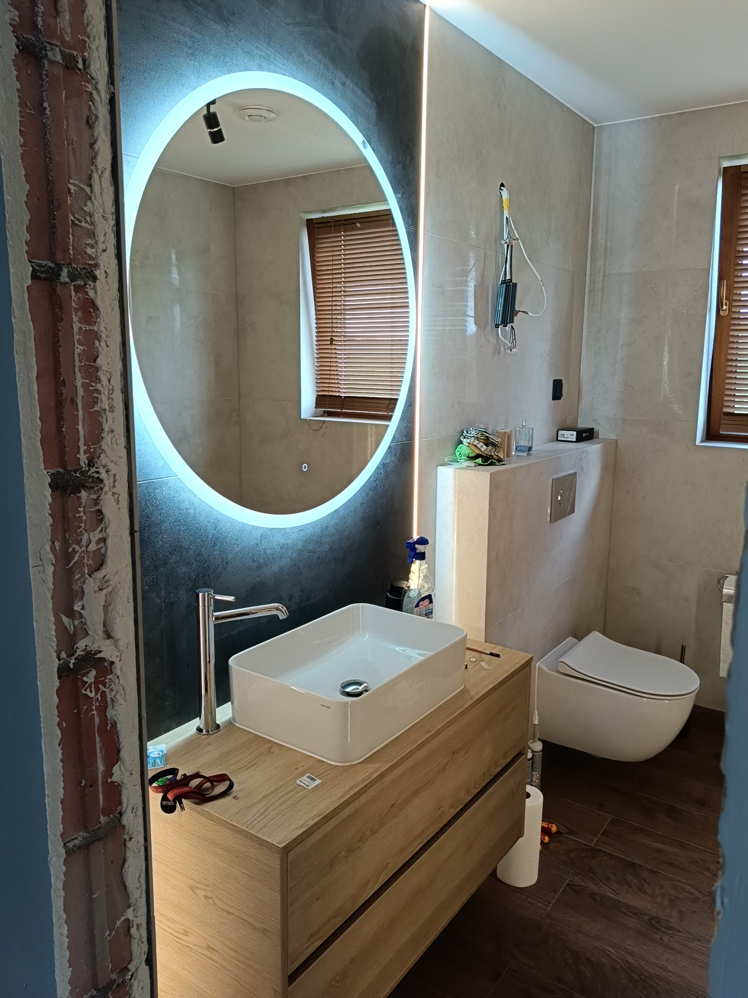 Nowoczesna łazienka w trakcie wykończenia: umywalka nablatowa, podświetlane lustro, wisząca toaleta, brązowe płytki imitujące drewno. Widoczne ślady prac remontowych.