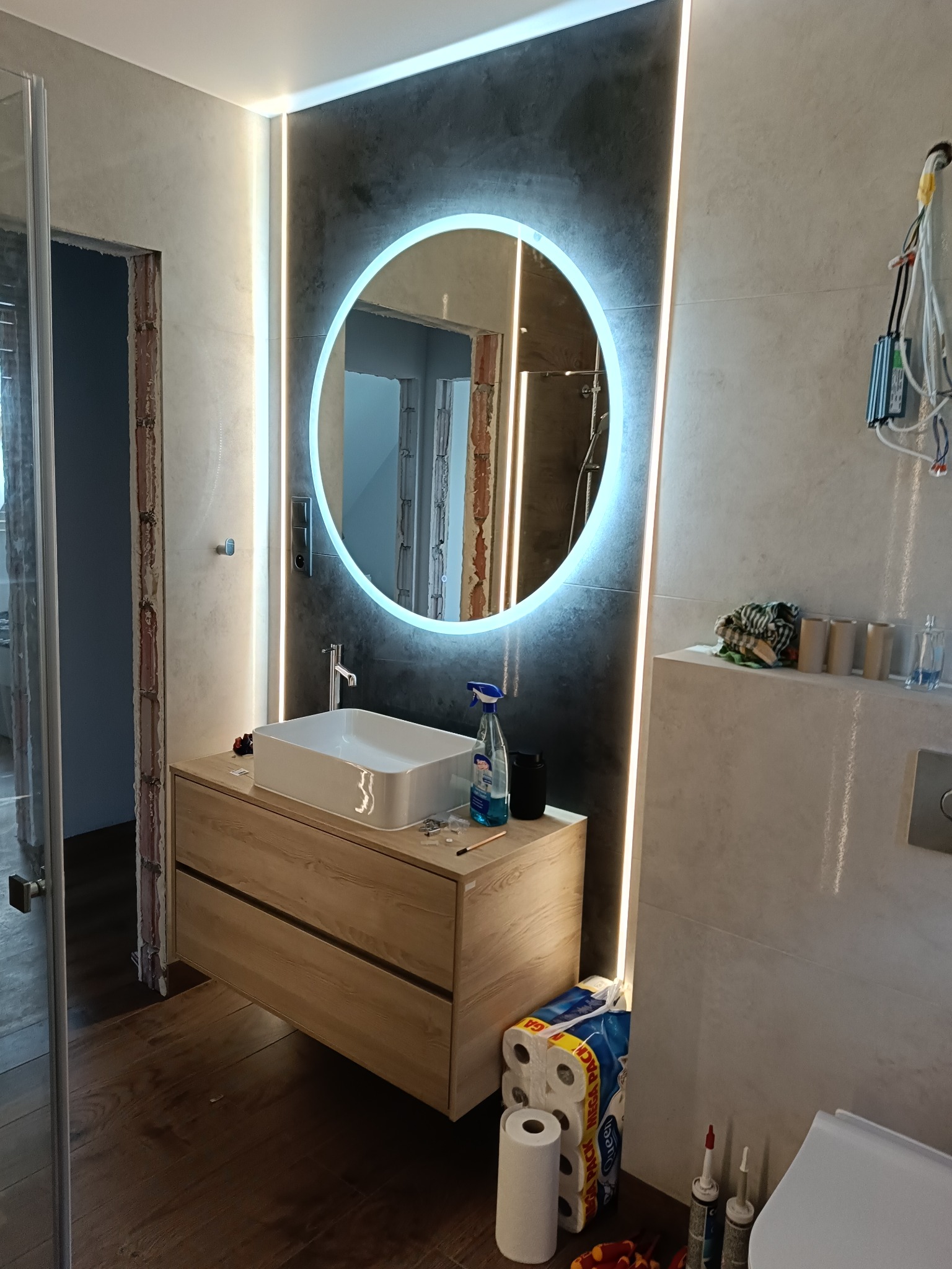 Nowoczesna łazienka w trakcie wykańczania: okrągłe lustro z podświetleniem LED, drewniana szafka pod umywalką i widoczne materiały budowlane.