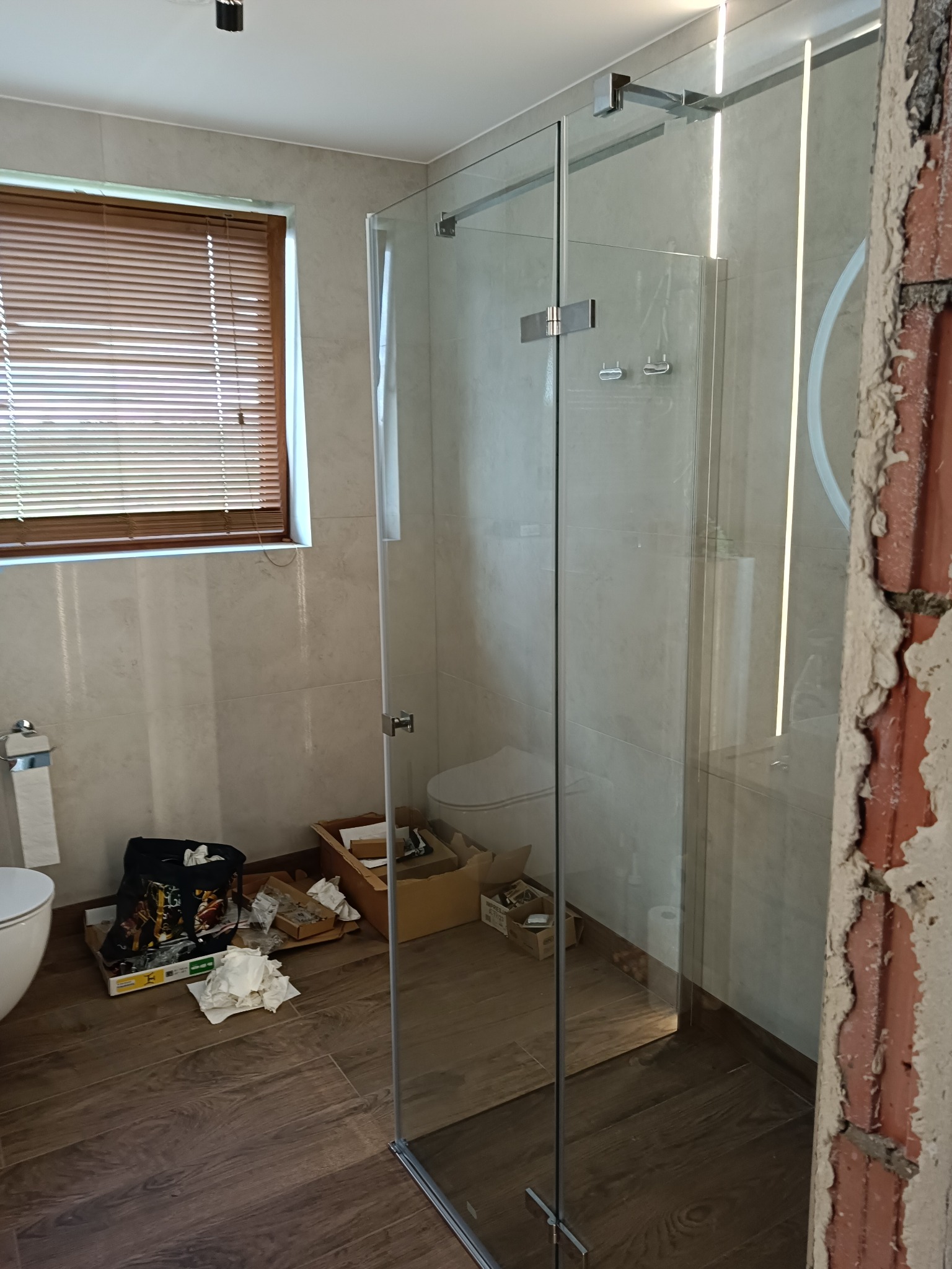Szklana kabina prysznicowa z chromowanymi okuciami w łazience z drewnopodobną podłogą i beżowymi płytkami, widok na okno z żaluzjami i fragment ściany w trakcie wykończenia.