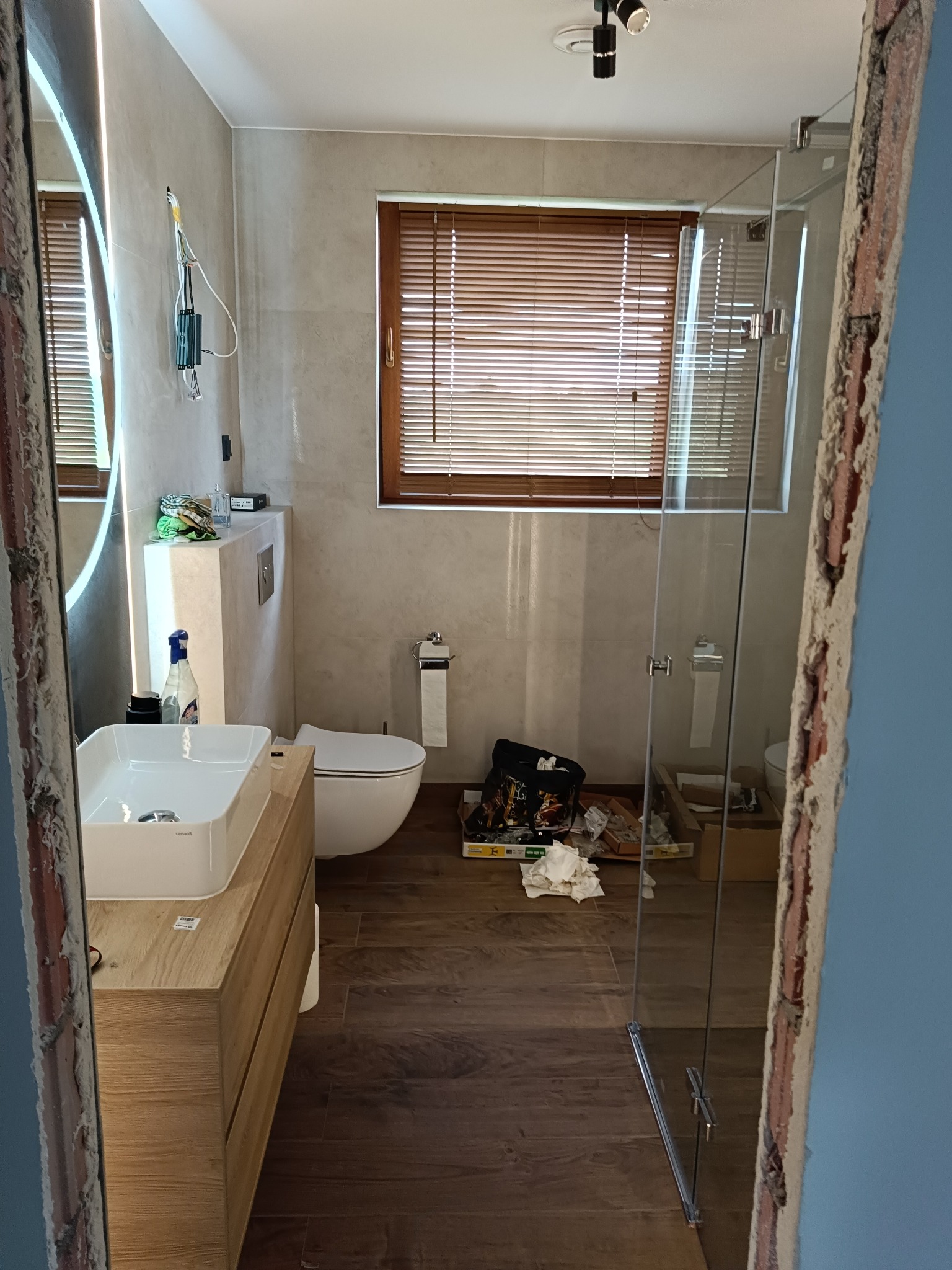 Wykończona łazienka z drewnianą podłogą i szafką pod umywalką, widok na kabinę prysznicową i okno z żaluzjami. Fragment ściany w stanie surowym.