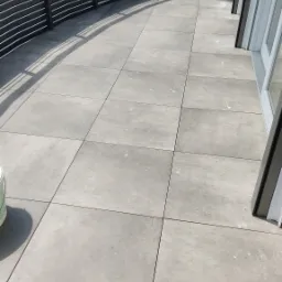 Wyłożony szarymi płytkami taras z metalową balustradą i widocznymi drzwiami balkonowymi, uchwycony w dziennym świetle z lekkiego ukosu.