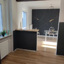 Nowoczesne wnętrze z czarną ścianą akcentową z motywem złotych żył, drewnianą podłogą i barkiem oddzielającym kuchnię od jadalni, widoczne dekoracje świąteczne.