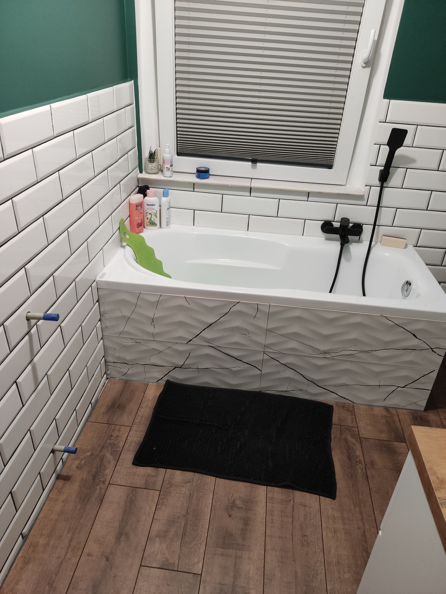 Biała wanna obudowana płytkami z motywem pęknięć, czarny dywanik łazienkowy i płytki imitujące drewno na podłodze w łazience w Człuchowie.