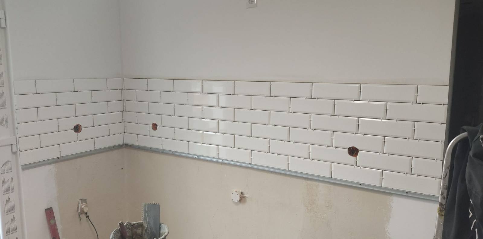 Białe, prostokątne płytki ułożone na ścianie do połowy wysokości, z widocznymi otworami na gniazdka elektryczne, metalowa listwa pod płytkami, wiadro i narzędzia do układania płytek widoczne...