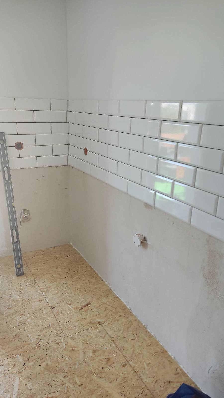 Ściana w trakcie układania białych, prostokątnych płytek, widoczny poziomica przy ścianie, surowa ściana poniżej płytek i płyta OSB na podłodze.