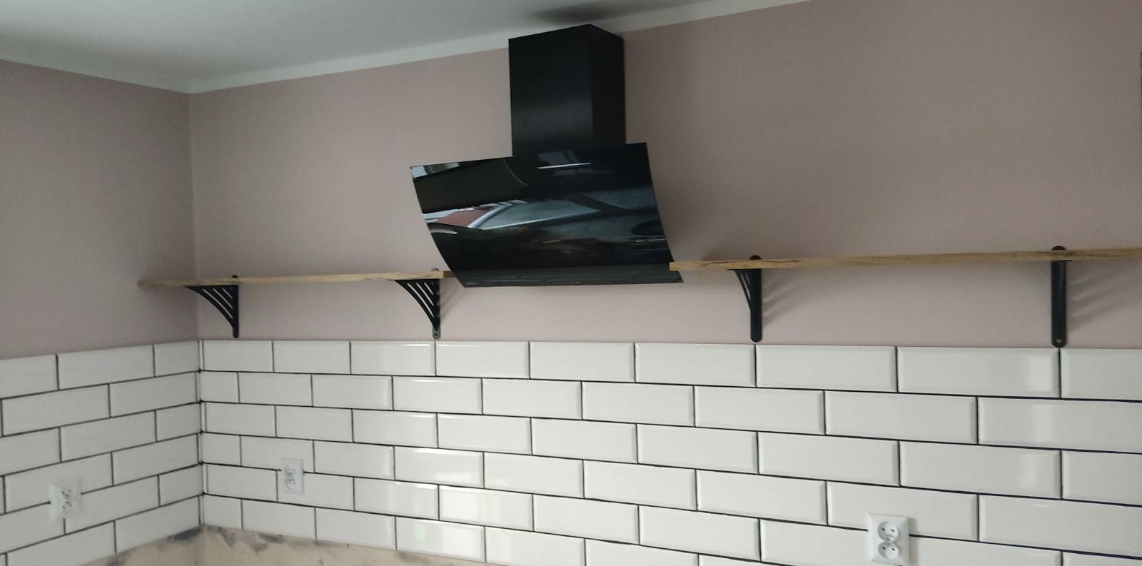Świeżo ułożone, białe, prostokątne płytki z ciemną fugą na ścianie w kuchni, z widocznym fragmentem ściany pomalowanej na jasny róż, czarnym okapem i surową, drewnianą półką na czarnych wspornikach.