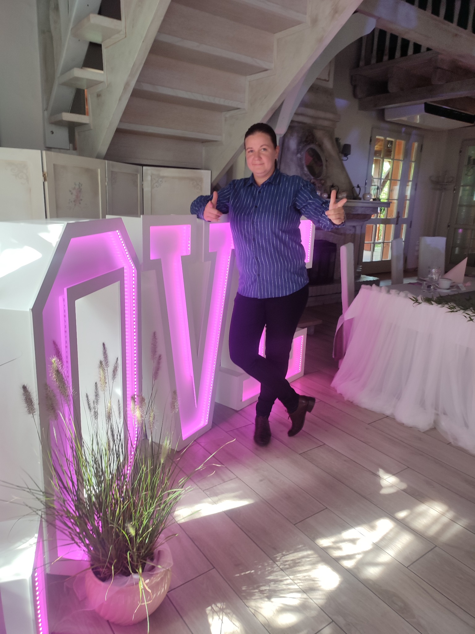 Kobieta opiera się o białe, podświetlane na różowo litery 'LOVE' w rustykalnym wnętrzu z drewnianymi schodami i stołem udekorowanym tiulem.