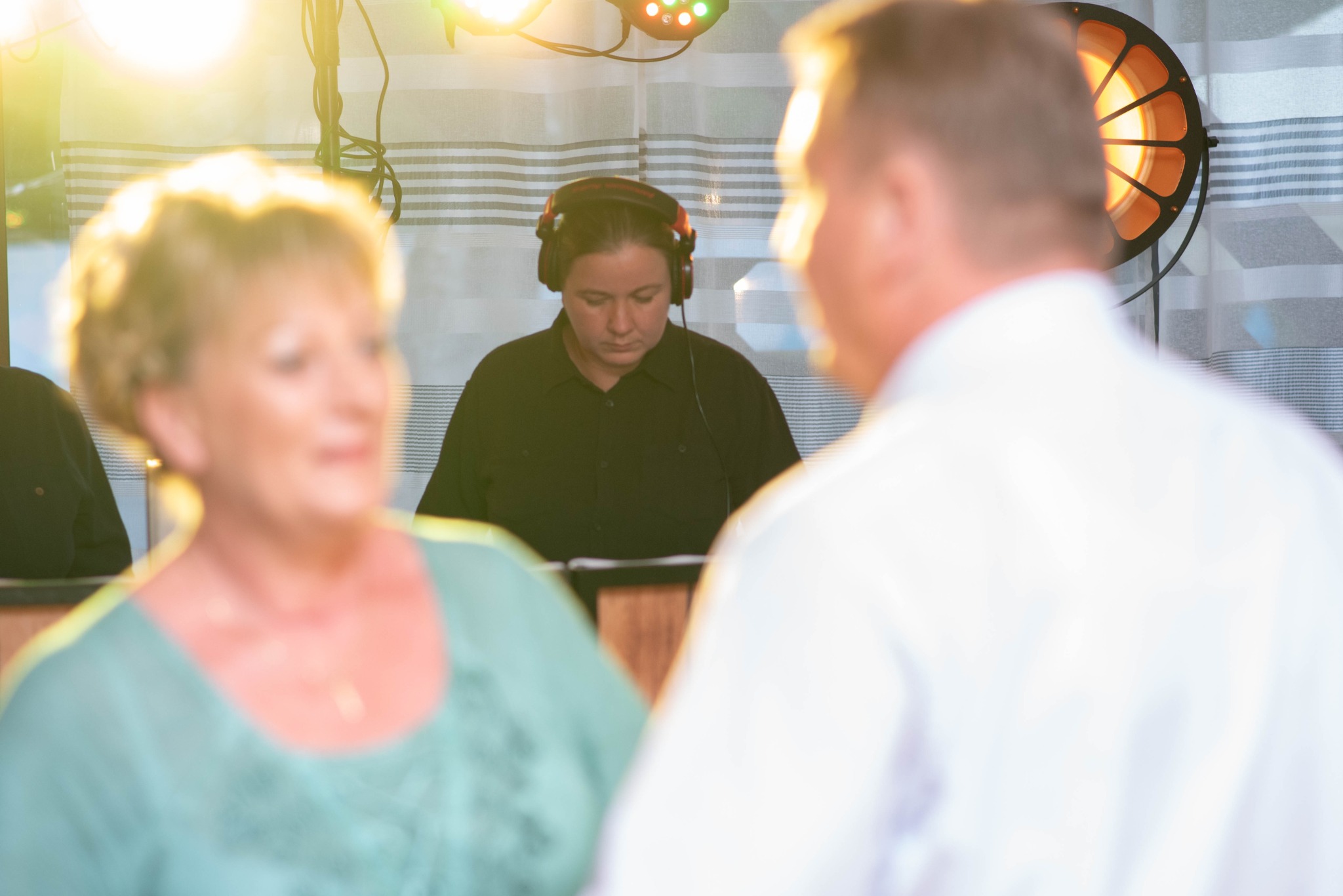 Kobieta DJ w słuchawkach, skupiona na pracy, oświetlenie sceniczne w tle, para tańcząca na pierwszym planie.