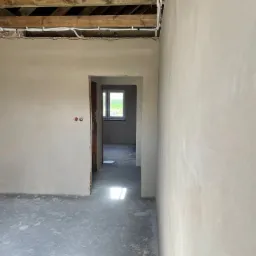 Surowe wnętrze z tynkami maszynowymi, widoczne drewniane belki stropowe i instalacja elektryczna, w tle okno z widokiem na zieleń.