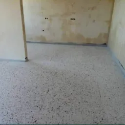 Pokój w trakcie remontu, z nową, jasną posadzką z drobnymi, ciemnymi plamkami, zabezpieczoną folią przy ścianach, które wymagają jeszcze wykończenia.