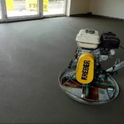 Świeżo wylana, szara posadzka jastrychowa, wygładzana mechaniczną zacieraczką marki Kreber, widoczne okna i ściany w surowym stanie.