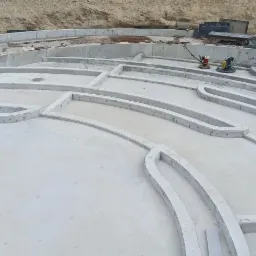 Widok z góry na betonowe fundamenty o nietypowym, organicznym kształcie, z widocznymi elementami zbrojenia i sprzętem budowlanym na obrzeżach terenu.