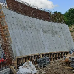 Betonowa ściana oporowa w trakcie budowy z widocznym zbrojeniem, szalunkiem i elementami konstrukcyjnymi, na tle nieba z chmurami.