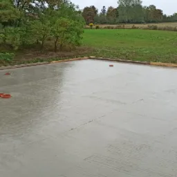 Świeżo wylana, mokra wylewka betonowa na budowie, widoczne pomarańczowe znaczniki i drewniane szalunki obwodowe, w tle zielone pole i drzewa pod pochmurnym niebem.