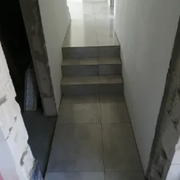 Szare, matowe płytki podłogowe ułożone na schodach i korytarzu w trakcie remontu, widoczne surowe ściany i materiały budowlane.