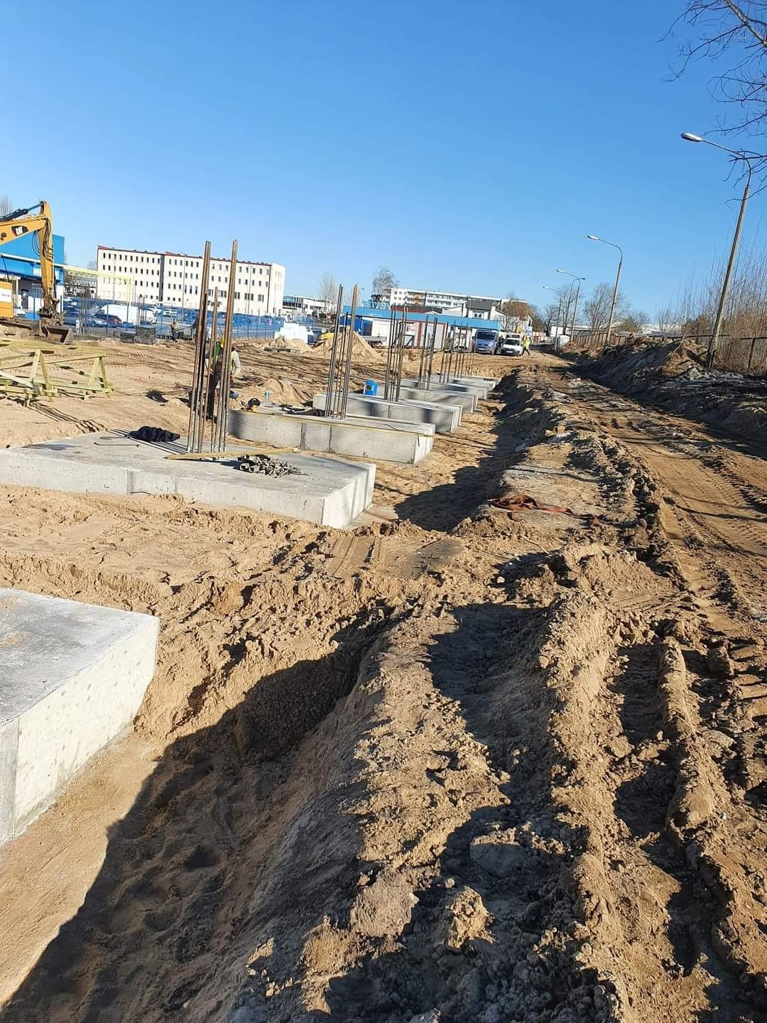 Fundamenty betonowe z wystającymi prętami zbrojeniowymi na placu budowy, widoczne ślady opon na piasku i budynek w tle.