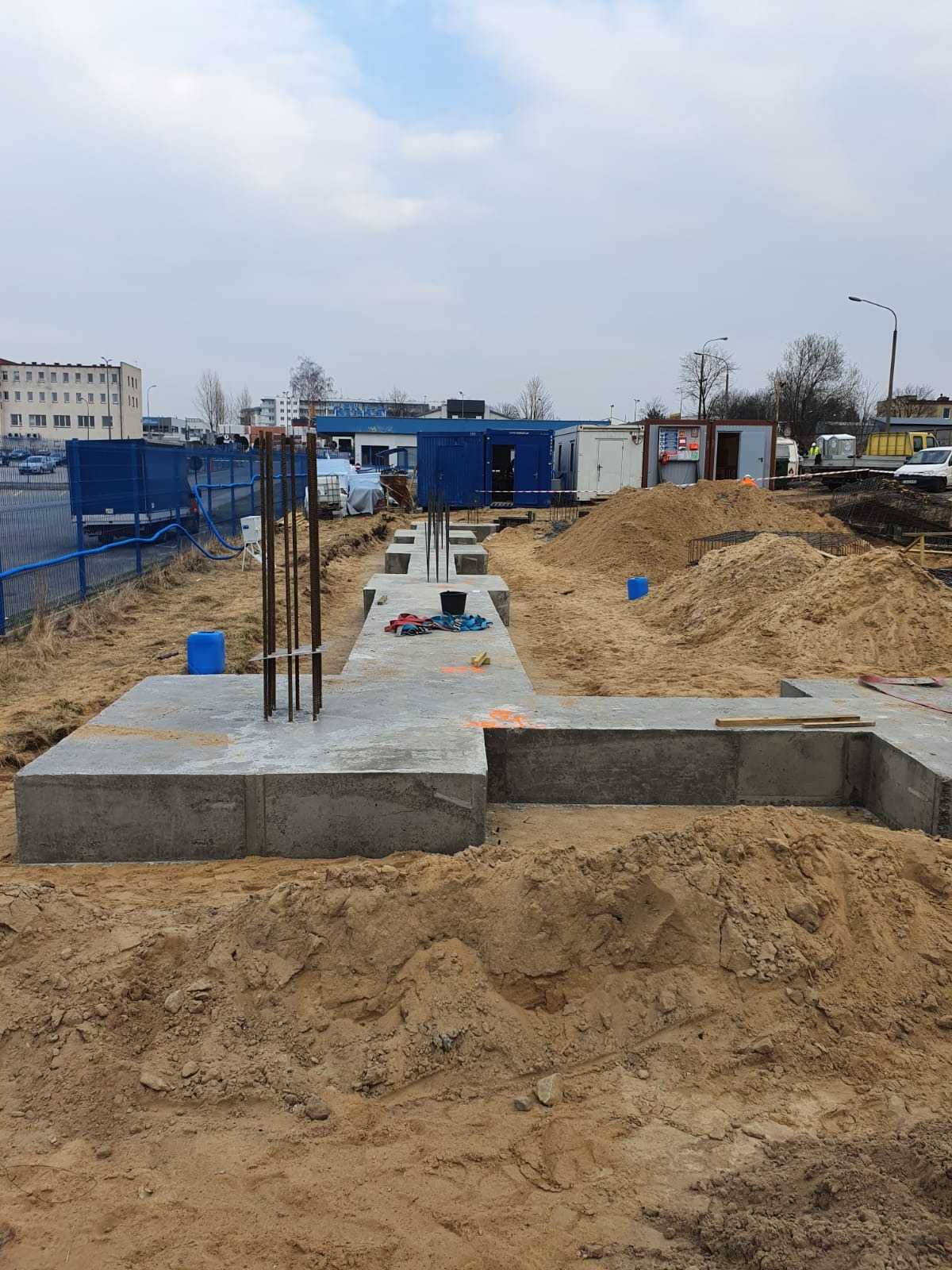 Betonowe fundamenty w trakcie budowy, widoczne zbrojenia, szalunki, teren budowy z piaskiem i kontenerami w tle.