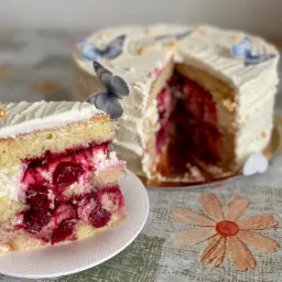 Kawałek tortu wiśniowego z białym kremem i złotymi kuleczkami na białym talerzyku, w tle tort udekorowany motylami.