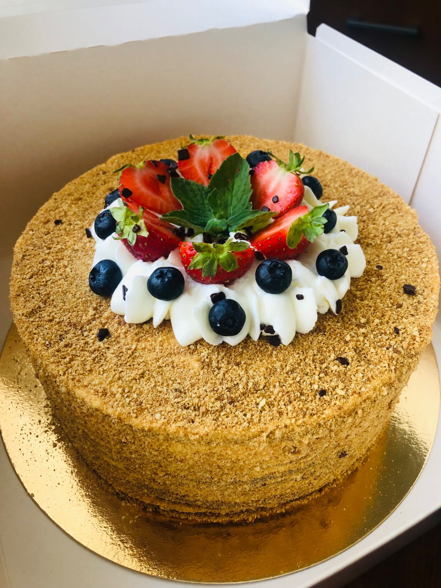 Tort miodowy z dekoracją z bitej śmietany, truskawek, borówek i listków mięty, umieszczony w kartonowym pudełku.