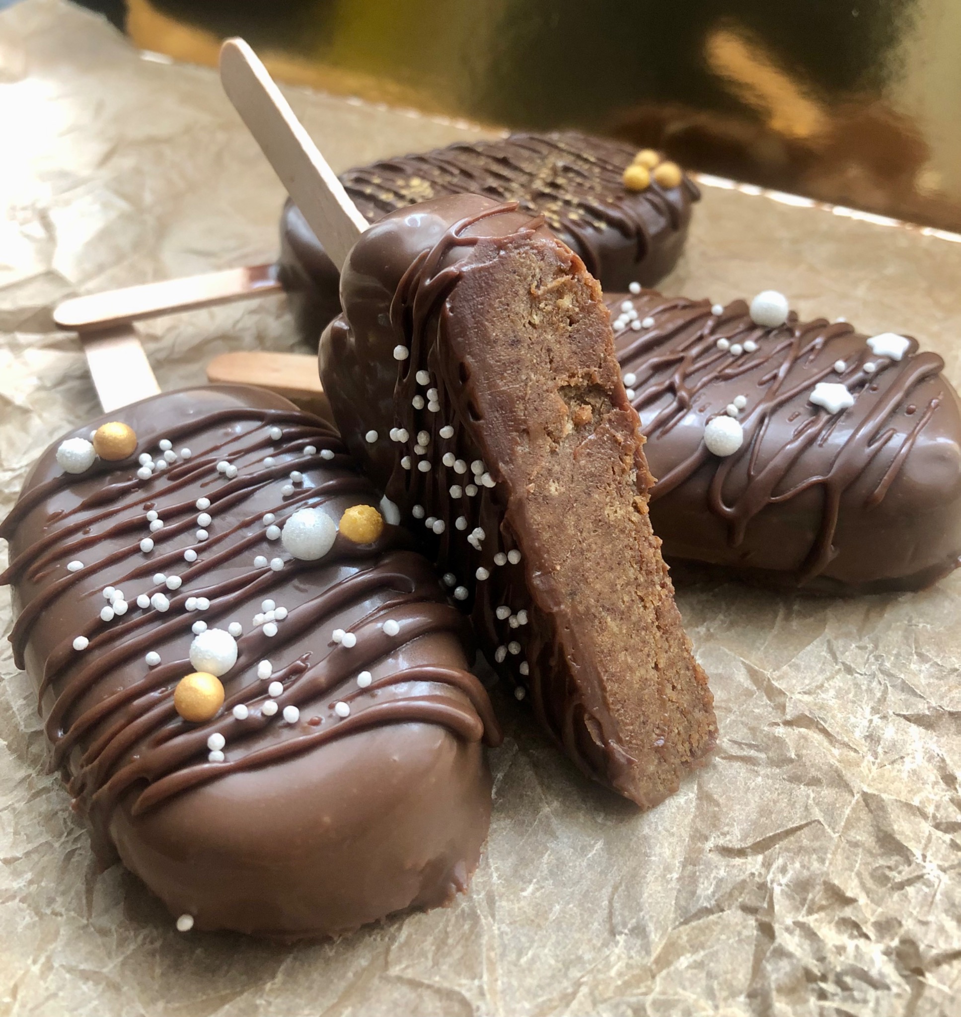 Cztery cake pops na patyku, oblane czekoladą i udekorowane posypką, jeden przekrojony, ukazujący strukturę ciasta, leżą na zmiętej papierowej powierzchni.