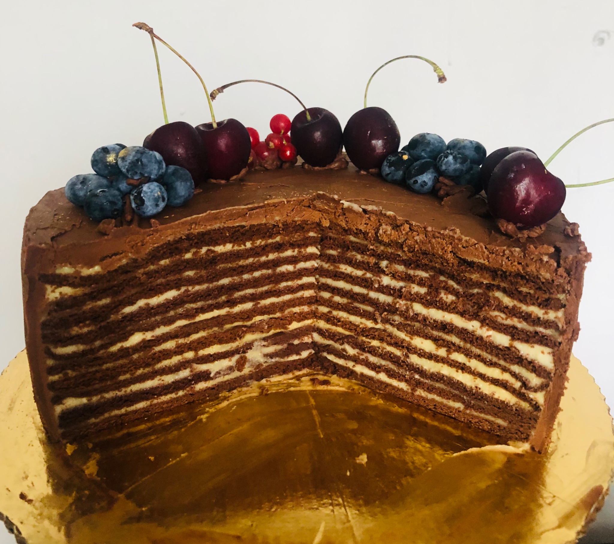 Przekrój wielowarstwowego tortu czekoladowego z kremem, udekorowanego wiśniami, borówkami i czerwoną porzeczką na złotym podkładzie.