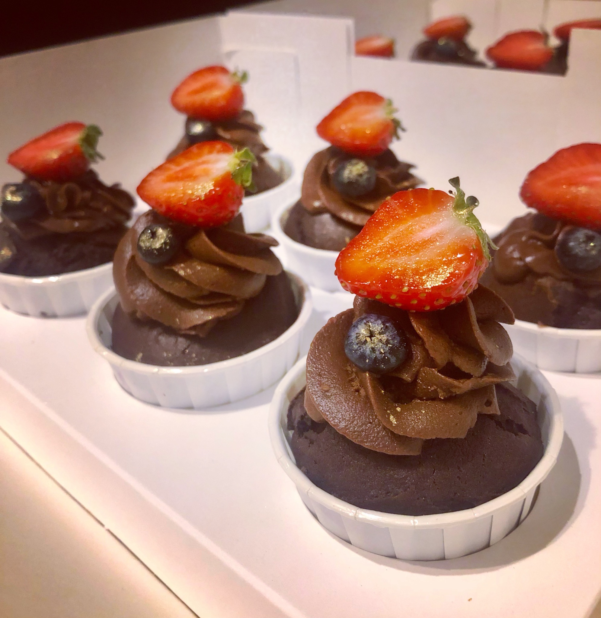 Rząd czekoladowych babeczek w białych papilotkach, każda udekorowana kremem czekoladowym, połówką truskawki i pojedynczą borówką z drobinkami złota.