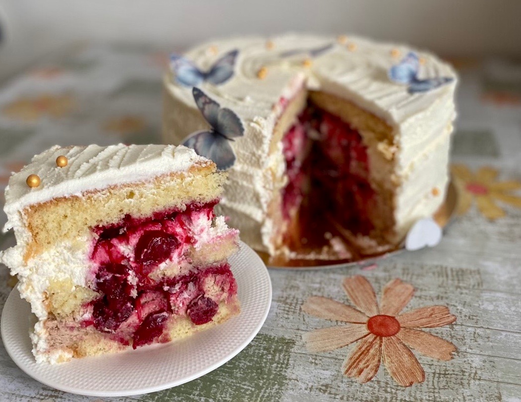 Kawałek tortu wiśniowego z białym kremem i złotymi kuleczkami na białym talerzyku, w tle tort udekorowany motylami.