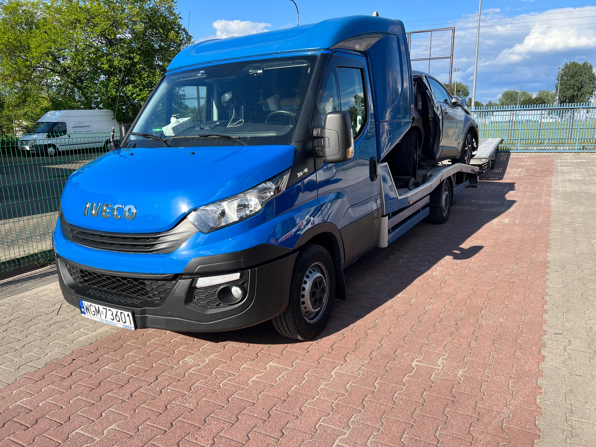 Niebieski samochód marki Iveco z lawetą przewozi uszkodzony, szary samochód osobowy z otwartymi drzwiami na tle ogrodzenia i zieleni.