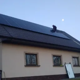 Montaż paneli słonecznych na dachu domu jednorodzinnego o zmierzchu, widoczny pracownik w kombinezonie i drabina oparta o ścianę. Część paneli już zamontowana, jeden panel oparty o ścianę budynku.