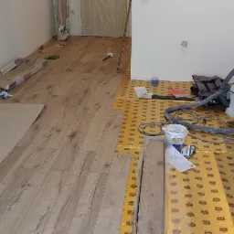 Układanie paneli podłogowych: widok na częściowo ułożoną podłogę z paneli drewnopodobnych, z widocznymi narzędziami, odkurzaczem przemysłowym Festool, podkładem pod panele i listwami...