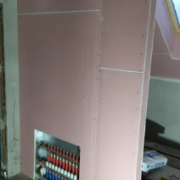 Montaż płyt gipsowo-kartonowych z wycięciem na rozdzielacz ogrzewania podłogowego; widoczne łączenia płyt i wkręty.