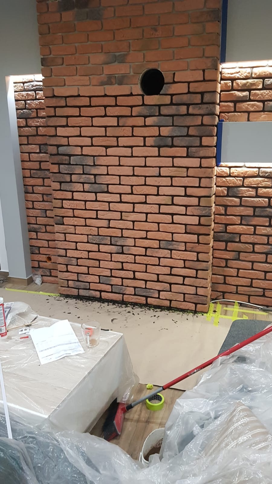 Wykończona ściana z cegieł klinkierowych z otworem, widoczne narzędzia i resztki materiałów budowlanych po remoncie.