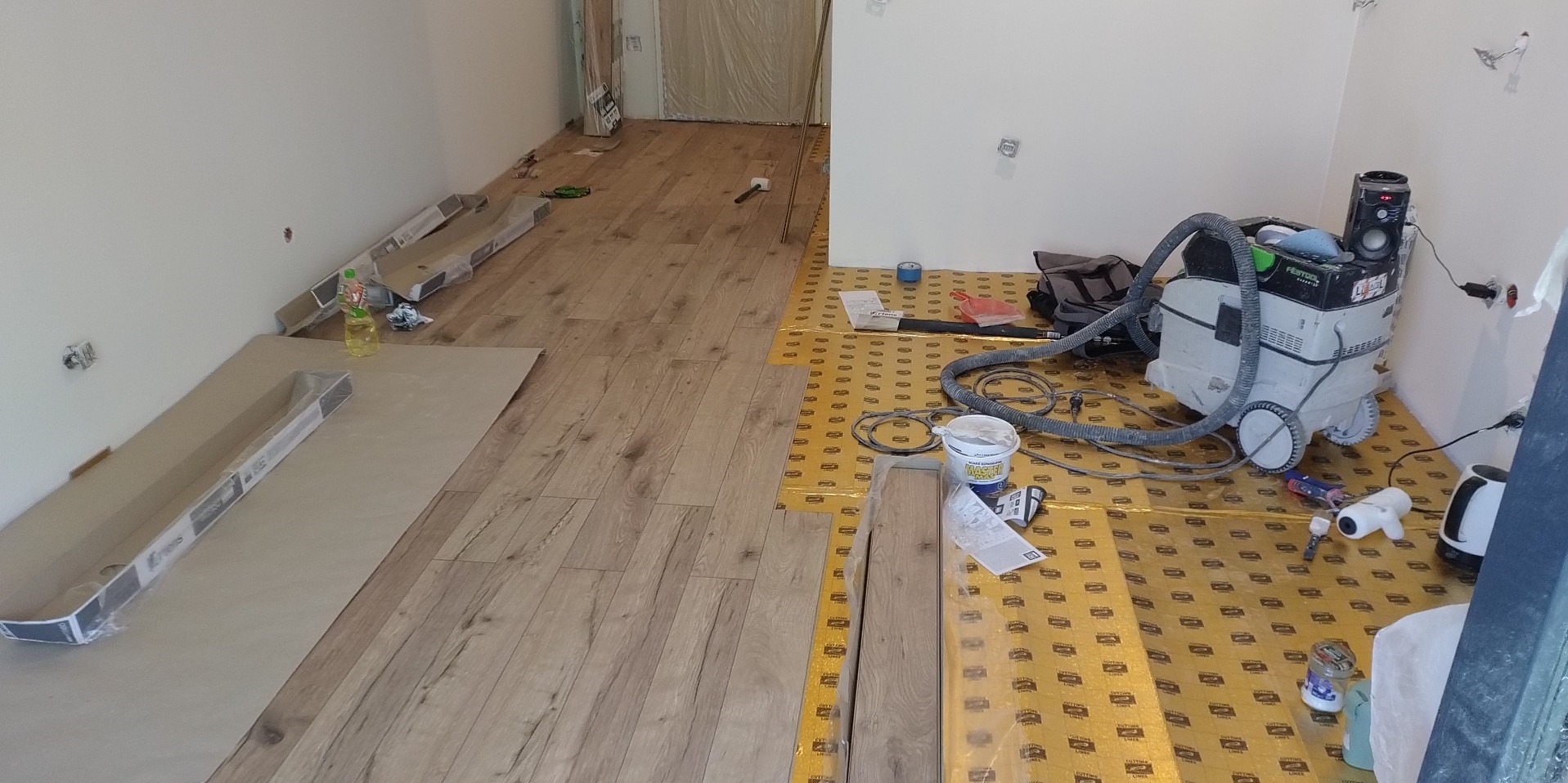 Układanie paneli podłogowych: widok na częściowo ułożoną podłogę z paneli drewnopodobnych, z widocznymi narzędziami, odkurzaczem przemysłowym Festool, podkładem pod panele i listwami...