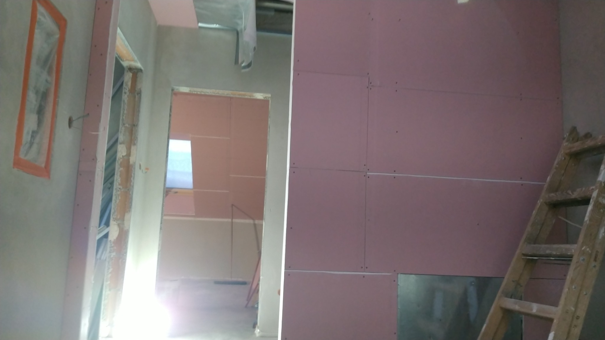 Montaż płyt gipsowo-kartonowych na ścianach w trakcie remontu, widoczna drabina oparta o ścianę, zabezpieczone futryny, surowy stan wykończenia.