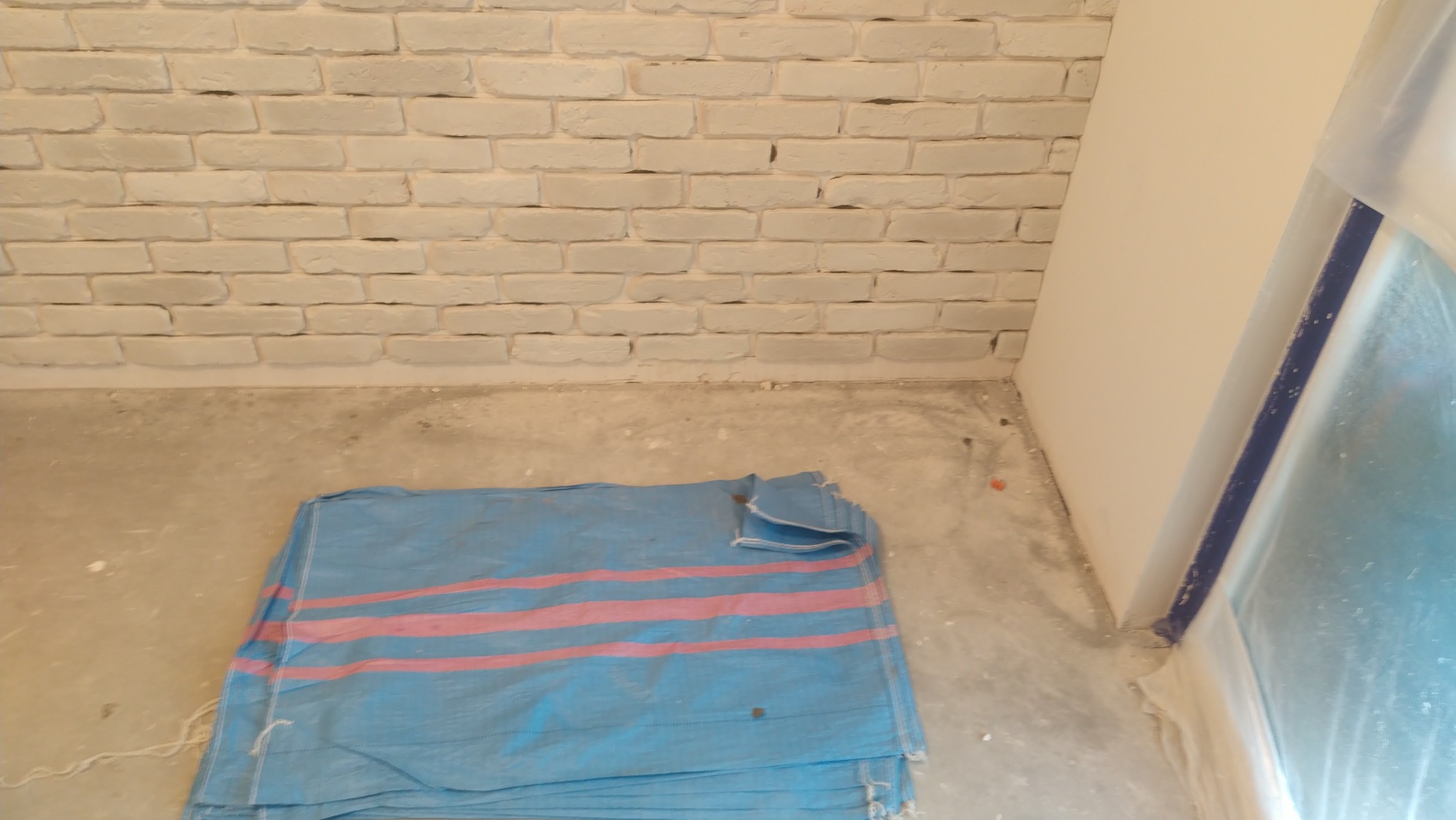 Świeżo pomalowana biała ściana z cegieł, zabezpieczona folią okienna i podłoga przykryta plandeką w niebiesko-różowe pasy