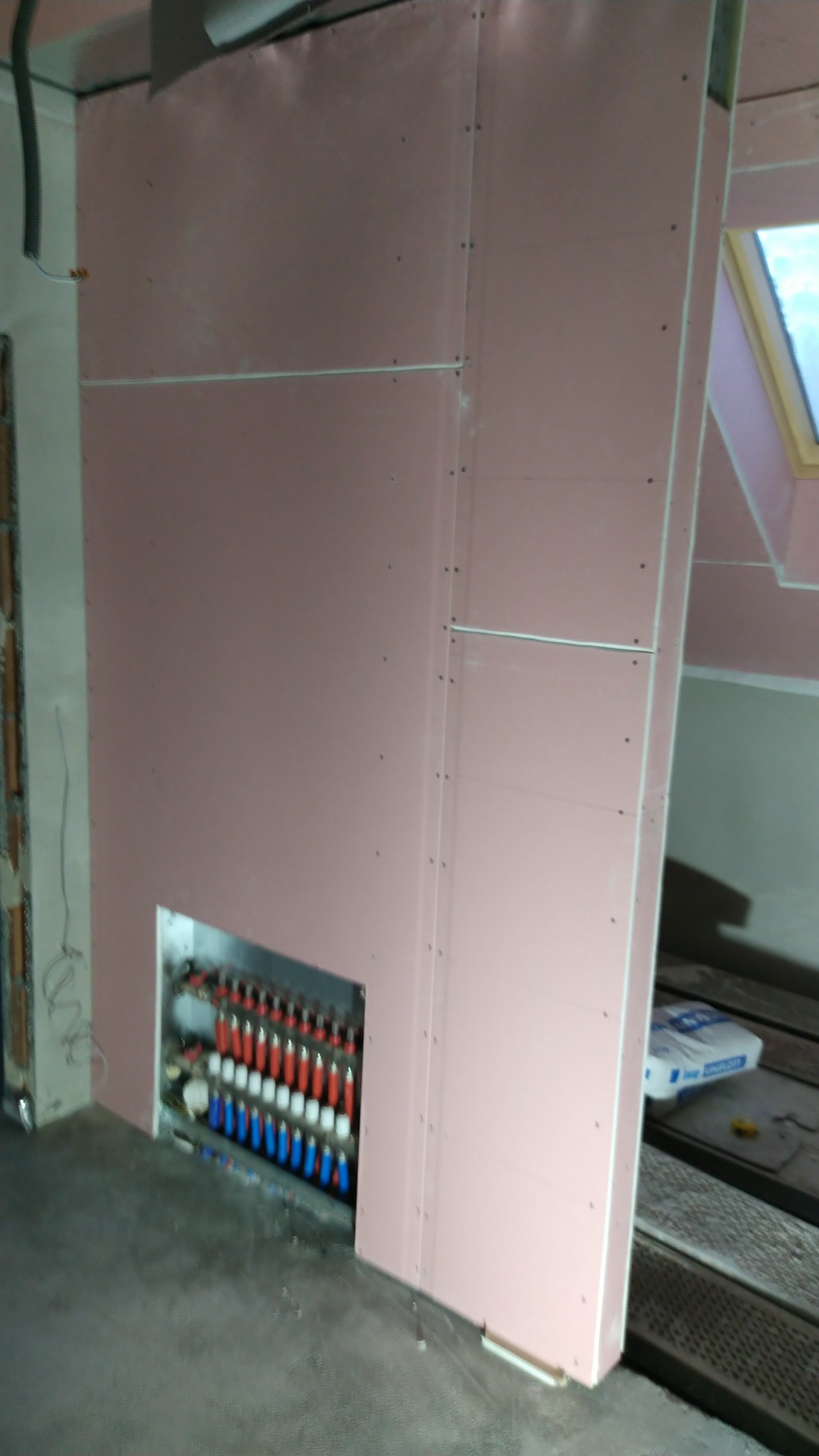 Montaż płyt gipsowo-kartonowych z wycięciem na rozdzielacz ogrzewania podłogowego; widoczne łączenia płyt i wkręty.