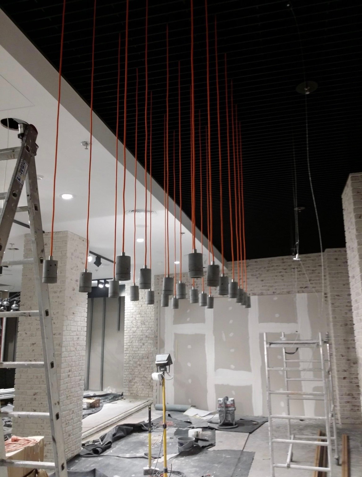Montaż sufitowych lamp wiszących z pomarańczowymi kablami w surowym wnętrzu sklepu, widoczna drabina i przygotowane ściany.
