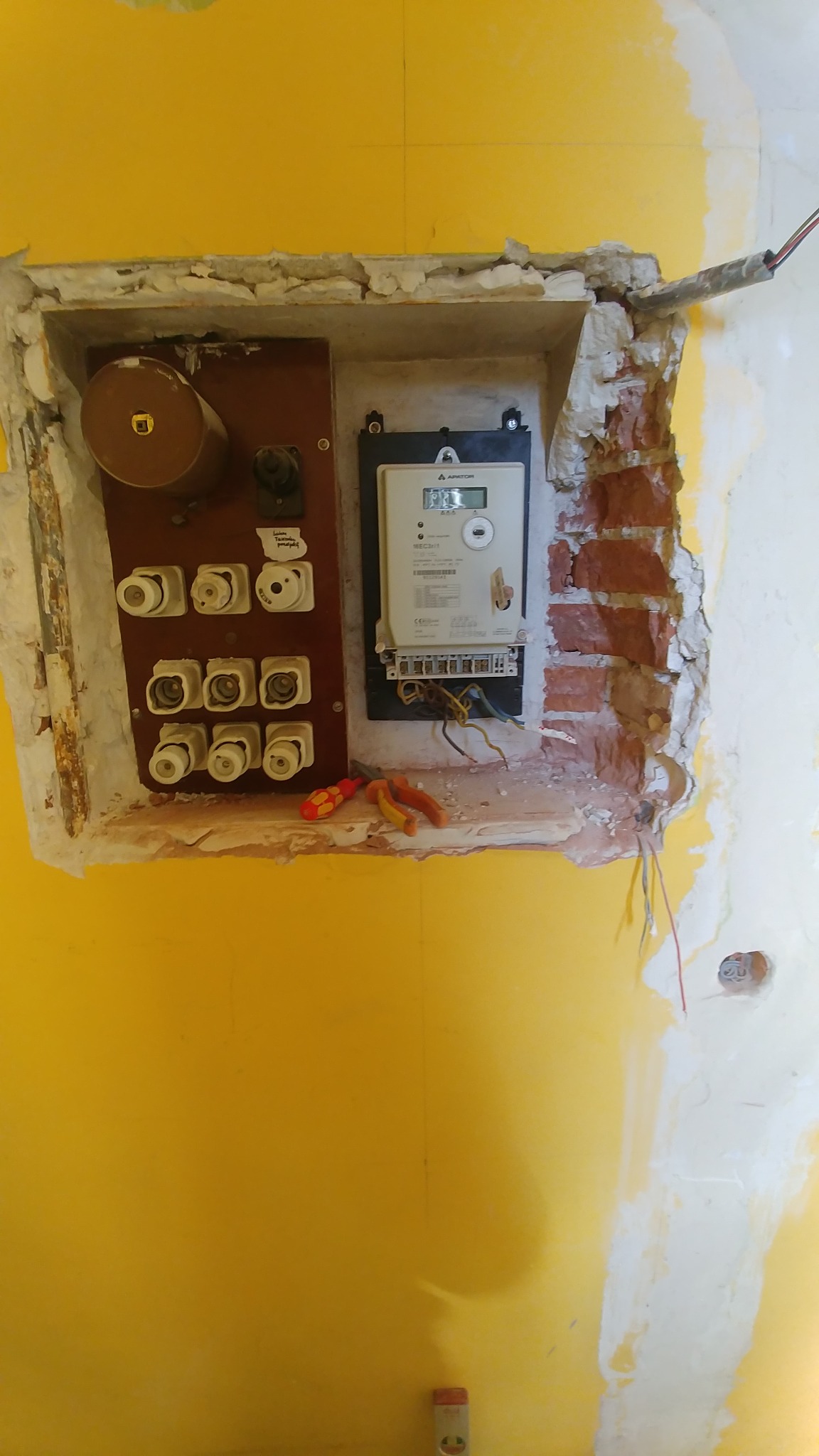 Stara tablica bezpiecznikowa z bezpiecznikami topikowymi i licznik energii elektrycznej w trakcie demontażu, widoczne narzędzia i odsłonięta cegła w ścianie.