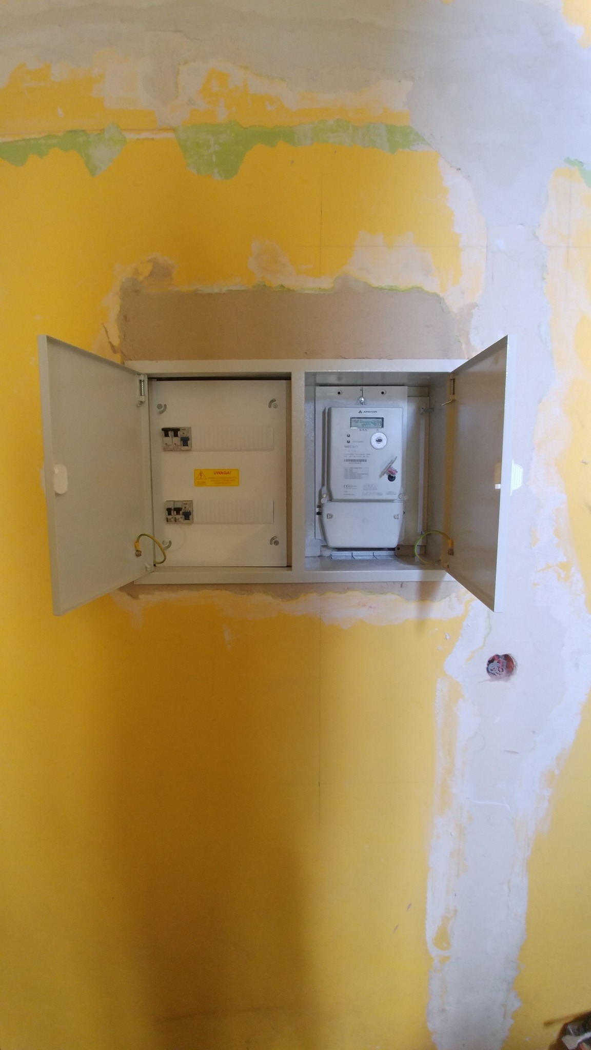 Otwarta skrzynka z bezpiecznikami i licznikiem elektrycznym zamontowana na żółtej ścianie w trakcie remontu, widoczne ślady po malowaniu.