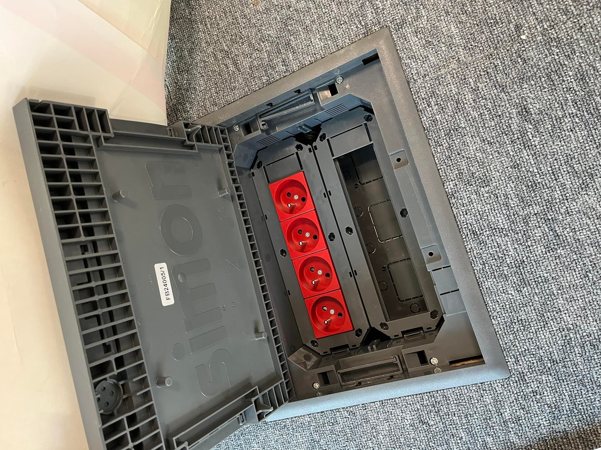 Otwarta podłogowa puszka instalacyjna z czterema czerwonymi gniazdami elektrycznymi, widoczny numer seryjny FB24005/1 na pokrywie, zamontowana w szarej wykładzinie dywanowej.