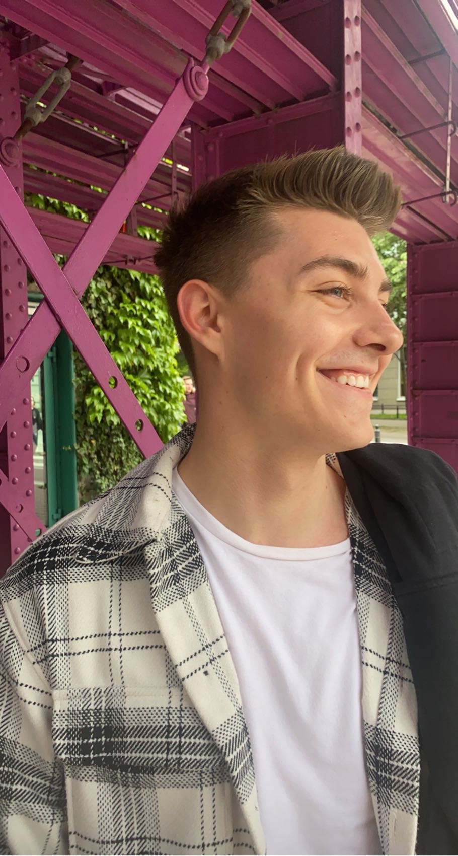 Uśmiechnięty młody mężczyzna w koszuli w kratę, stojący na tle fioletowej konstrukcji i zielonej roślinności.