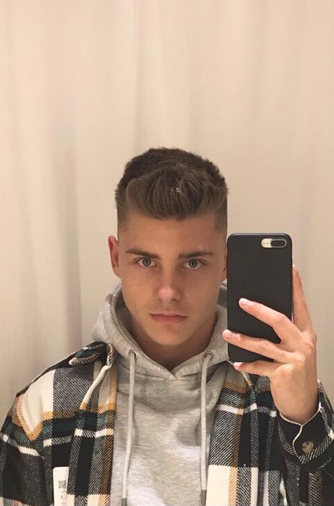 Młody mężczyzna z krótkimi włosami robi selfie trzymając czarny telefon, ubrany w szarą bluzę z kapturem i flanelową koszulę w kratę na tle jasnej ściany.