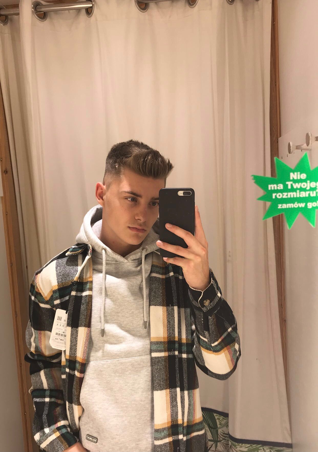 Młody mężczyzna robi selfie w przymierzalni, ubrany w szarą bluzę z kapturem i flanelową koszulę w kratę; na ścianie naklejka z informacją o dostępności rozmiarów.