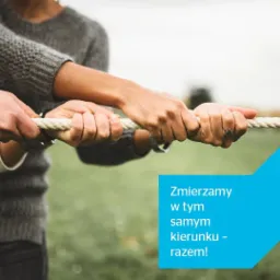 Dłonie zaciskają się na linie podczas przeciągania, symbolizując współpracę i dążenie do wspólnego celu, z hasłem 'Zmierzamy w tym samym kierunku - razem!' na niebieskim tle.