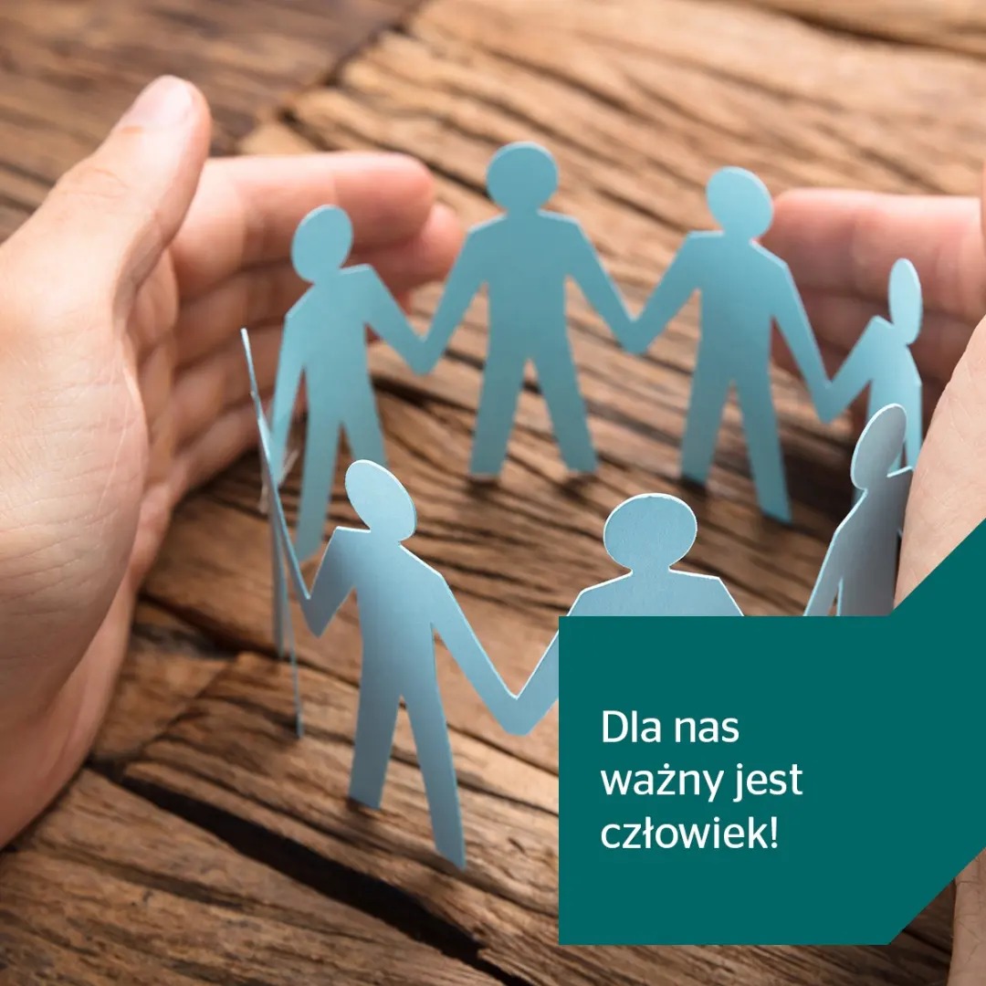 Dłonie otaczają krąg papierowych ludzików na drewnianym tle, symbolizując ochronę i wsparcie, z hasłem 'Dla nas ważny jest człowiek!'