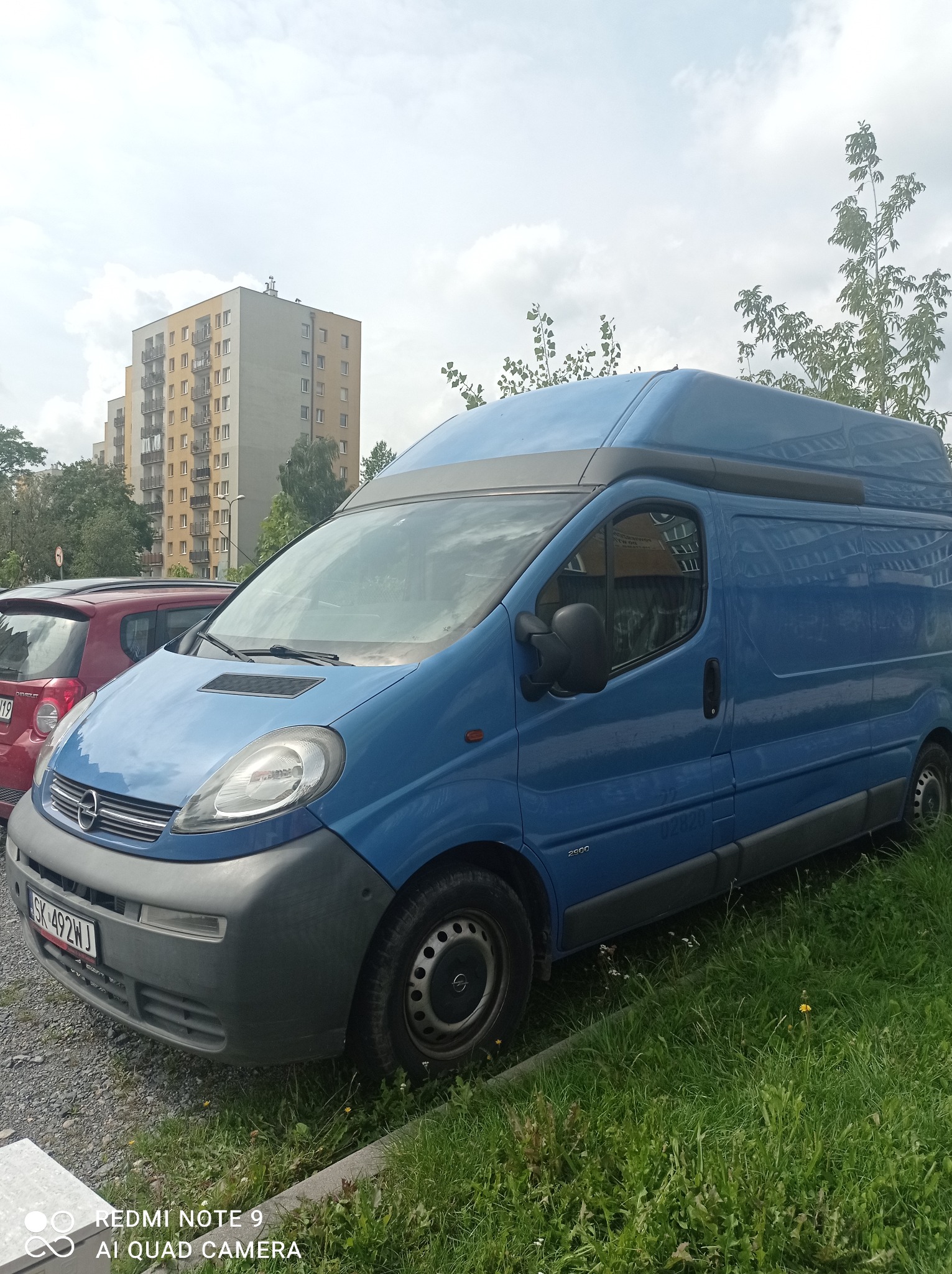 Niebieski bus Opel Vivaro z podwyższonym dachem zaparkowany na trawie przed blokiem mieszkalnym, widoczna tablica rejestracyjna SK 492WJ.