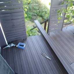 M...K - Montaż kompozytowej deski tarasowej w Wałbrzychu: widok z góry na częściowo ukończony taras z ciemnobrązowych desek, w tle zieleń ogrodu, widoczne narzędzia pracy: wiertarka, pojemnik ze śrubami...